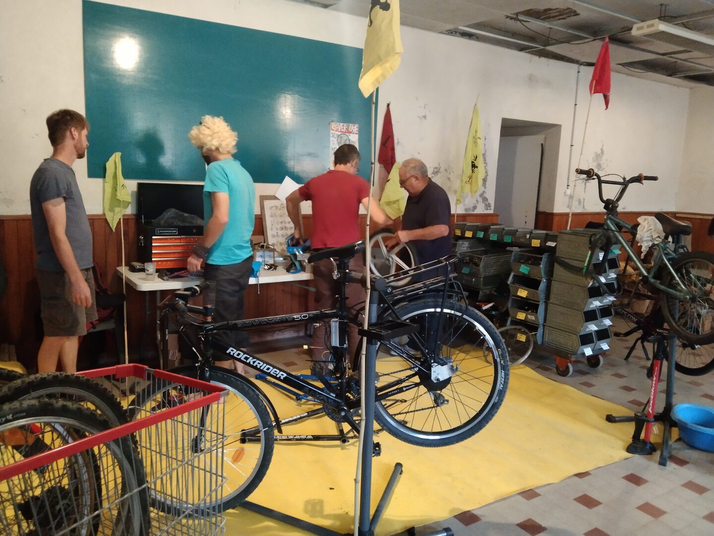 3 hommes bricolant dans une ancienne salle de classe aménagée en atelier éphémère de réparation de vélos. L'un d'eux porte une perruque blonde. Au premier plan, un vélo noir en cours de réparation.